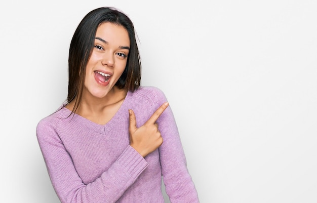 Junges hispanisches Mädchen in lässiger Kleidung, fröhlich, mit einem Lächeln im Gesicht, das mit der Hand und dem Finger zur Seite zeigt, mit einem glücklichen und natürlichen Gesichtsausdruck