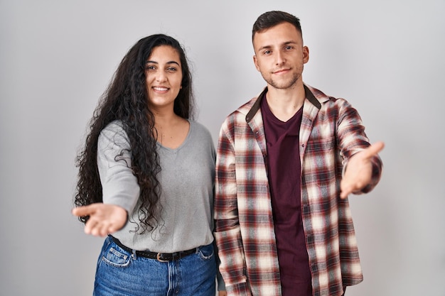 Kostenloses Foto junges hispanisches paar, das vor weißem hintergrund steht und fröhlich lächelt und seine handfläche anbietet, um hilfe und akzeptanz zu geben