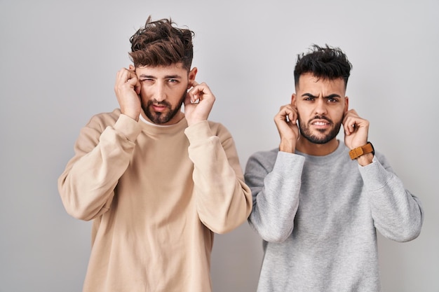 Junges homosexuelles Paar, das vor weißem Hintergrund steht und die Ohren mit den Fingern bedeckt, mit genervtem Ausdruck für den Lärm lauter Musik. Gehörloses Konzept.