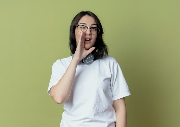 Junges hübsches kaukasisches Mädchen mit Brille und Kopfhörern am Hals, das etwas flüstert und schaut