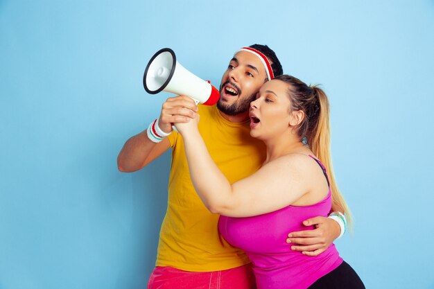 Junges hübsches kaukasisches Paar im hellen Kleidertraining auf blauem Hintergrund Konzept des Sports, der menschlichen Gefühle, des Ausdrucks, des gesunden Lebensstils, der Beziehung, der Familie. Der Umsatz. In Mundfrieden zusammenrufen.