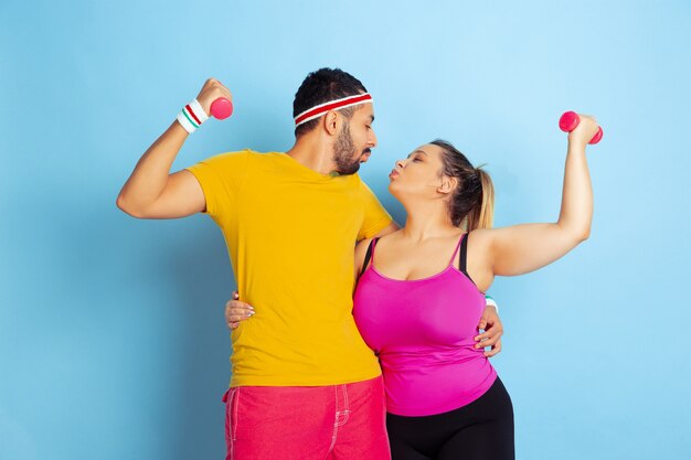 Junges hübsches kaukasisches Paar im hellen Kleidertraining auf blauem Hintergrund Konzept des Sports, der menschlichen Gefühle, des Ausdrucks, des gesunden Lebensstils, der Beziehung, der Familie. Training mit Gewichten, viel Spaß.