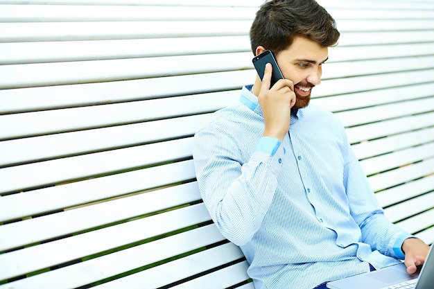 Junges hübsches lächelndes Geschäftsmannmodell, das auf der Parkbank im lässigen Hipster-Stoff sitzt, der auf Handy spricht