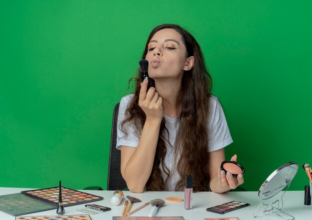 Junges hübsches Mädchen, das am Make-up-Tisch mit Make-up-Werkzeugen sitzt, die erröten Bürste halten und erröten, die Pinsel und gestikulierenden Kuss lokalisiert auf grünem Hintergrund betrachten