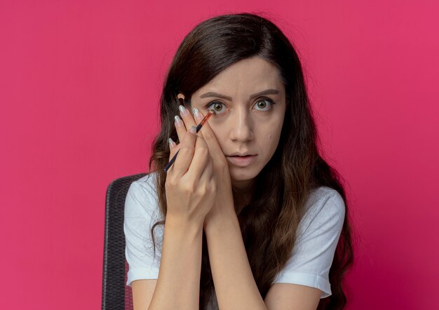 Junges hübsches mädchen, das am make-up-tisch mit make-up-werkzeugen sitzt, die gesicht berühren und lidschatten mit make-up-pinsel anwenden, der kamera lokal auf purpurrotem hintergrund betrachtet