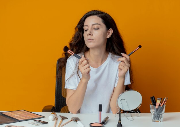 Junges hübsches Mädchen, das am Make-up-Tisch mit Make-up-Werkzeugen sitzt und Make-up-Pinsel lokalisiert auf orange Hintergrund hält