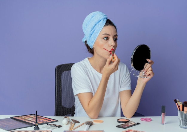 Junges hübsches Mädchen, das am Make-up-Tisch mit Make-up-Werkzeugen und mit Badetuch auf Kopf hält Spiegel hält und auf roten Lippenstift lokalisiert auf lila Hintergrund setzt