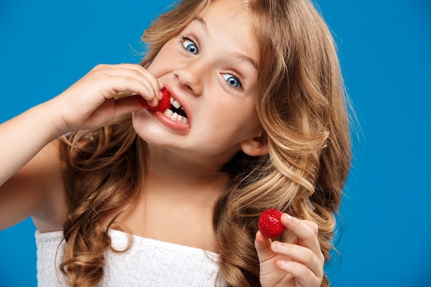 Junges hübsches Mädchen, das Erdbeere über blauer Wand isst