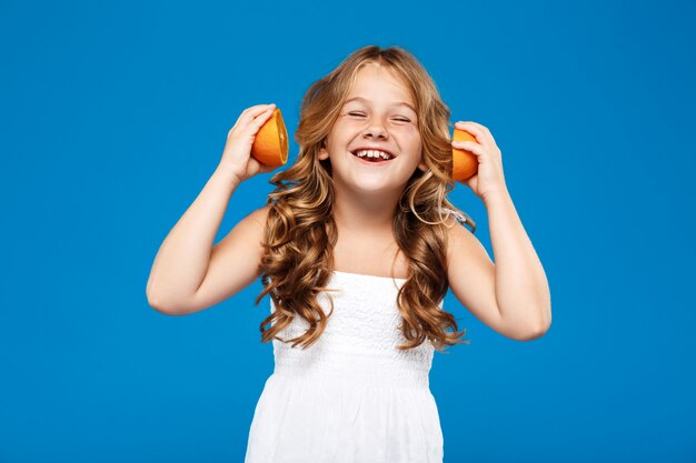 Junges hübsches Mädchen, das Orangen hält und über blaue Wand lächelt