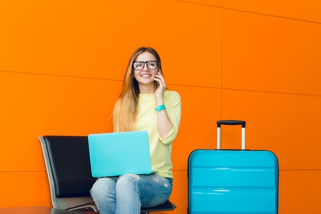 Junges hübsches Mädchen mit langen Haaren sitzt auf Stuhl auf orange Hintergrund. Sie trägt einen gelben Pullover, Jeans und eine Brille. Sie hat einen blauen Laptop auf den Knien und einen Koffer. Am Telefon sprechen.