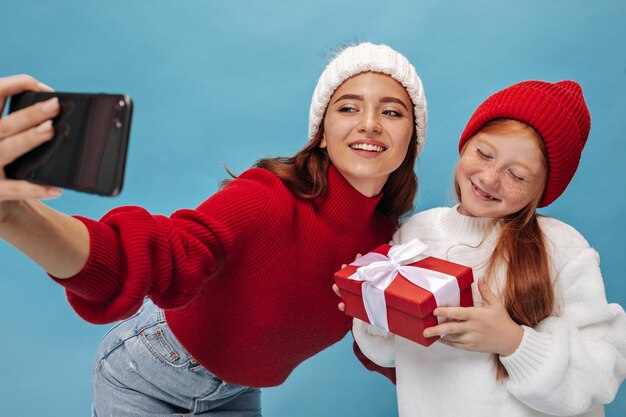 Junges hübsches Mädchen mit Sommersprossen in Hut und weißem Outfit hält rote Geschenkbox und macht Foto mit älterer stilvoller Schwester in hellem Pullover