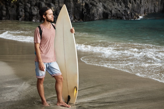 Junges hübsches männliches Modell, das mit Surfbrett auf exotischem Strand aufwirft