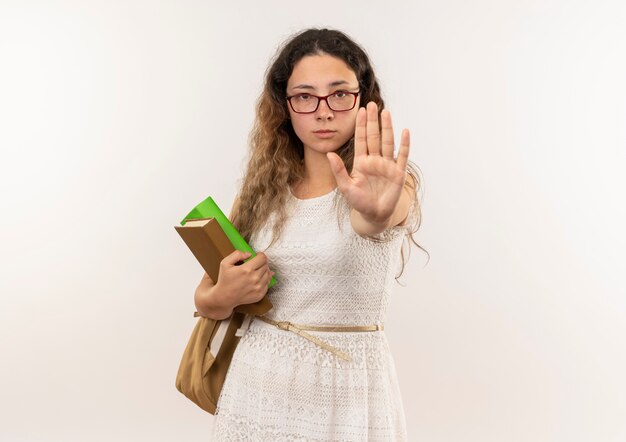 Junges hübsches Schulmädchen, das Brille und Rückentasche hält Bücher hält, die suchen und gestikulieren, stoppt an der Kamera lokalisiert auf weißem Hintergrund mit Kopienraum