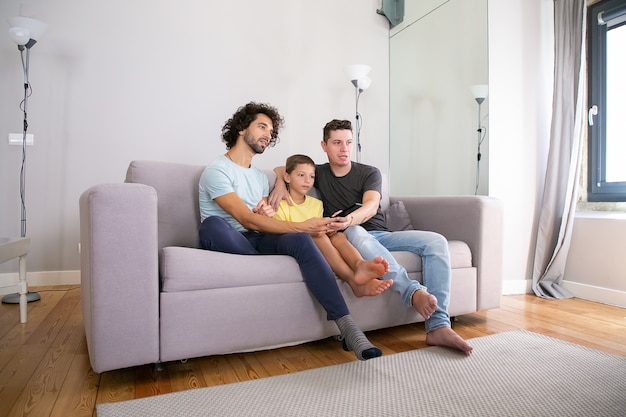 Junges hübsches schwules Paar und ihr Sohn, die Fernsehshow zu Hause schauen, auf Couch im Wohnzimmer sitzen, umarmen, Fernbedienung benutzen, wegschauen. Familien- und Home-Entertainment-Konzept