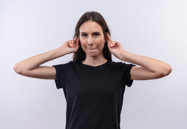 junges kaukasisches Mädchen, das schwarzes T-Shirt trägt, legte ihre Finger um Ohren auf isolierte weiße Wand