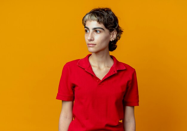 Junges kaukasisches Mädchen mit Pixie-Haarschnitt, der Seite lokalisiert auf orange Hintergrund mit Kopienraum betrachtet