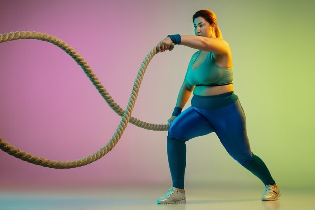 Junges kaukasisches plus size weibliches Model Training auf lila grüner Wand des Farbverlaufs im Neonlicht. Trainingsübungen mit Seilen machen. Konzept des Sports, gesunder Lebensstil, Körper positiv, Gleichheit.