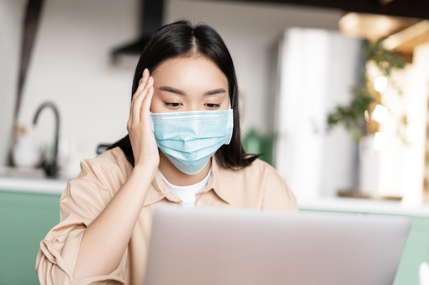 Junges krankes asiatisches Mädchen in medizinischer Gesichtsmaske mit Laptop, das von zu Hause aus in Quarantäne arbeitet