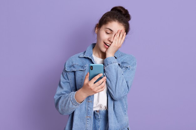 Junges lachendes brünettes Teenager-Mädchen mit Haarknoten, das Smartphone in Händen hält und die Hälfte des Gesichts mit Handfläche bedeckt
