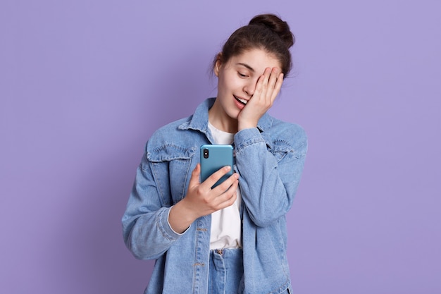 Junges lachendes brünettes Teenager-Mädchen mit Haarknoten, das Smartphone in Händen hält und die Hälfte des Gesichts mit Handfläche bedeckt