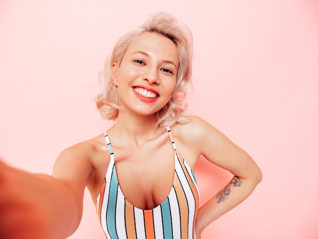 Junges lächelndes blondes Modell im bunten Badeanzug der Sommerbadebekleidung Sexy sorglose Frau, die Spaß hat und verrückt wird Weibliche Aufstellung nahe rosa Wand im Studio Fröhlich und glücklich Unter POV-Selfie