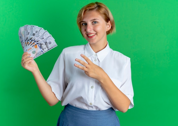 junges lächelndes blondes russisches Mädchen hält Geld und gestikuliert drei mit den Fingern