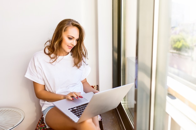 Junges lächelndes brünettes Mädchen sitzt auf modernem Stuhl nahe dem Fenster im hellen gemütlichen Raum zu Hause. Sie arbeitet in entspannter Atmosphäre am Laptop