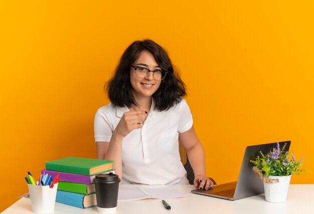 Junges lächelndes hübsches kaukasisches Schulmädchen, das Brille trägt, sitzt am Schreibtisch mit Schulwerkzeugen, die auf orange Raum mit Kopienraum isoliert werden