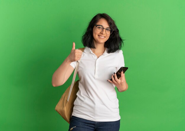 Junges lächelndes hübsches kaukasisches Schulmädchen, das Brille und Rückentasche Daumen hoch hält Telefon auf Grün mit Kopienraum hält