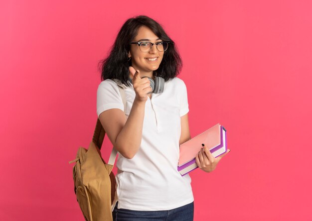 Junges lächelndes hübsches kaukasisches Schulmädchen, das Brille und Rückentasche mit Kopfhörern auf Halspunkten an der Kamera trägt, hält Bücher auf Rosa mit Kopienraum