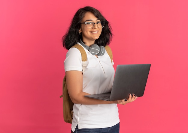 Junges lächelndes hübsches kaukasisches Schulmädchen mit Kopfhörern am Hals, das Brille und Rückentasche trägt, hält Laptop auf Rosa mit Kopienraum