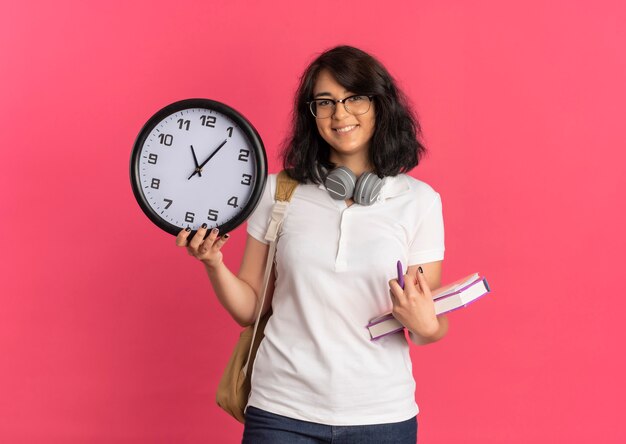 Junges lächelndes hübsches kaukasisches Schulmädchen mit Kopfhörern am Hals, das Brille und Rückentasche trägt, hält Uhr und Bücher auf Rosa mit Kopienraum