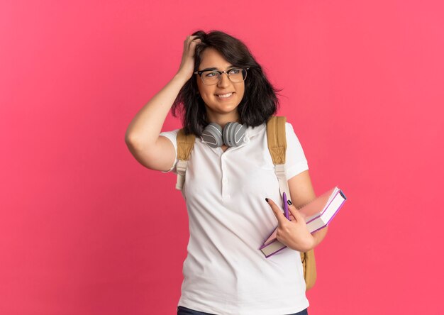 Junges lächelndes hübsches kaukasisches Schulmädchen mit Kopfhörern am Hals, das Brille und Rückentasche trägt, legt Hand auf Haar hält Buch schaut auf Seite auf Rosa mit Kopienraum