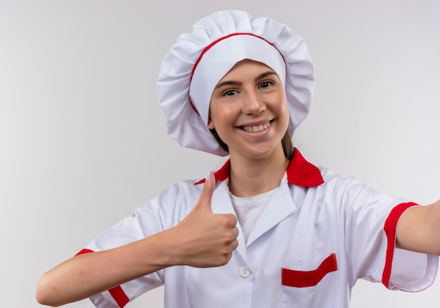Junges lächelndes kaukasisches Kochmädchen in der Kochuniform gibt vor, Kamera und Daumen auf Weiß mit Kopienraum zu halten