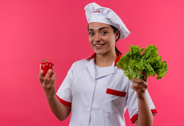 Junges lächelndes kaukasisches Kochmädchen in der Kochuniform hält roten Pfeffer und Salat lokalisiert auf rosa Wand mit Kopienraum