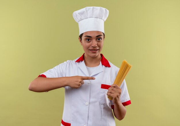Junges lächelndes kaukasisches Kochmädchen in der Kochuniform hält und zeigt auf Bündel von Spaghetti lokalisiert auf grüner Wand mit Kopienraum