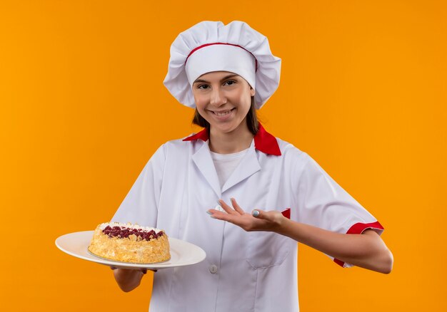 Junges lächelndes kaukasisches Kochmädchen in der Kochuniform hält und zeigt auf Kuchen auf Platte lokalisiert auf orange Raum mit Kopienraum