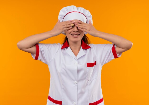 Junges lächelndes kaukasisches Kochmädchen in der Kochuniform schließt Augen mit den Händen, die auf orange Wand mit Kopienraum lokalisiert werden