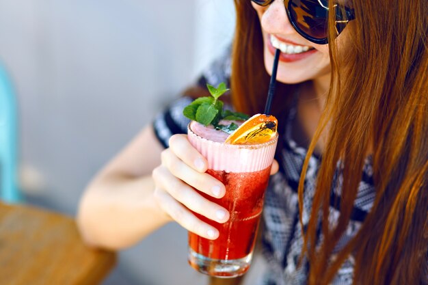 Junges lächelndes Mädchen, das leckeren süßen Cocktail, erstaunlichen entspannenden Tag, leckere Limonade, elegantes Kleid und Sonnenbrille, Außenterrasse trinkt.