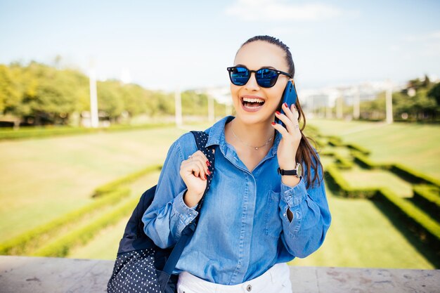 Junges lächelndes Mädchen in der Sonnenbrille, die in einem Park steht, während wegschaut und am Telefon spricht