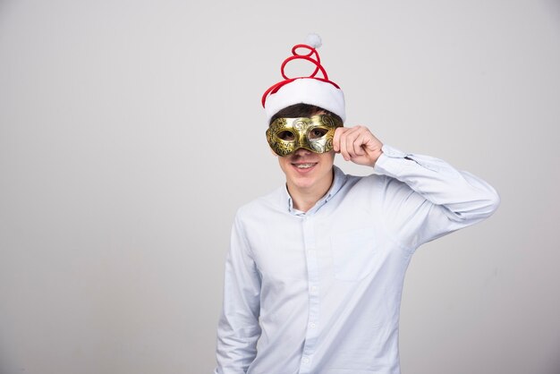 Junges lächelndes Mannmodell, das in Weihnachtsmütze mit Karnevalsmaske steht
