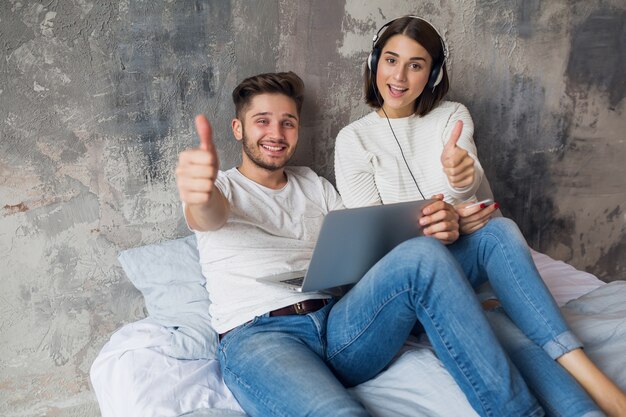 Junges lächelndes Paar, das auf Bett zu Hause im lässigen Outfit sitzt, Mann, der freiberuflich auf Laptop arbeitet, Frau, die Musik auf Kopfhörern hört, glückliche Zeit zusammen verbringt, positive Emotion, in der Kamera schauend