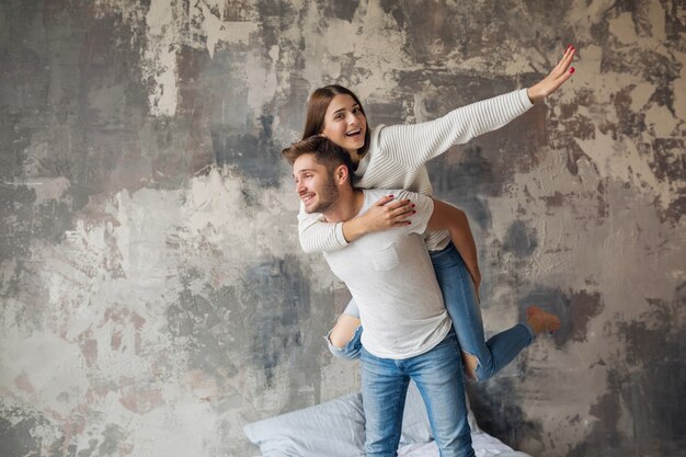 Junges lächelndes Paar, das auf Bett zu Hause im lässigen Outfit spielt, Mann und Frau, die Spaß zusammen haben, verrückte positive Emotion, glücklich, Hand hochhaltend