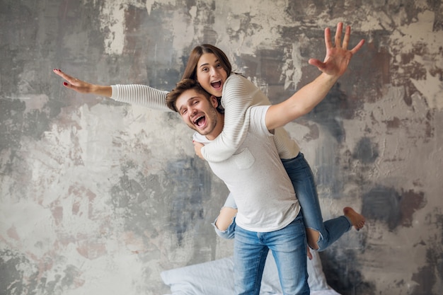 Junges lächelndes Paar, das auf Bett zu Hause im lässigen Outfit spielt, Mann und Frau, die Spaß zusammen haben, verrückte positive Emotion, glücklich, Hand hochhaltend