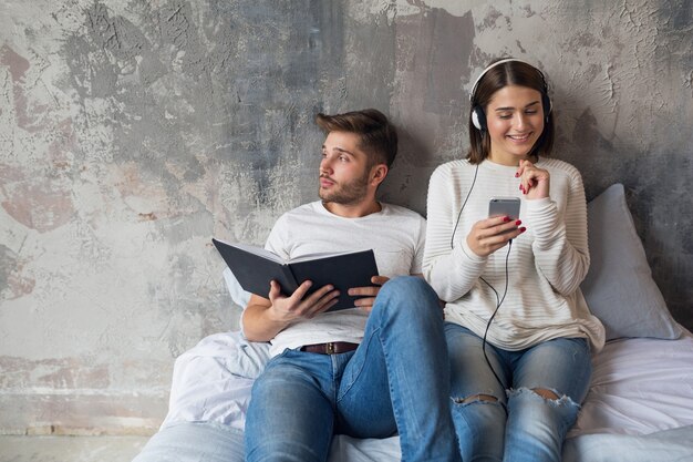 Junges lächelndes Paar, das auf dem Bett zu Hause im lässigen Outfit-Lesebuch sitzt, das Jeans trägt, Mann liest Buch, Frau, die Musik auf Kopfhörern hört, romantische Zeit zusammen verbringend