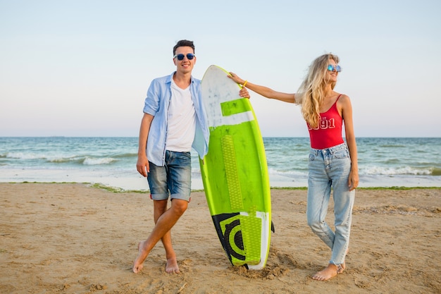 Junges lächelndes Paar, das Spaß am Strand hat, der mit Surfbrett aufwirft