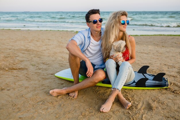 Junges lächelndes Paar, das Spaß am Strand hat, sitzt auf Sand mit Surfbrettern, die mit Hund spielen