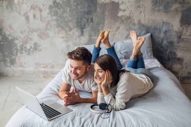 Junges lächelndes Paar, das zu Hause im lässigen Outfit auf Bett liegt und in Laptop, Mann und Frau schaut, die glückliche Zeit zusammen verbringen, entspannend
