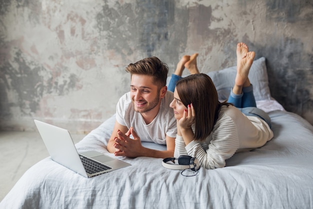 Junges lächelndes Paar, das zu Hause im lässigen Outfit auf Bett liegt und in Laptop, Mann und Frau schaut, die glückliche Zeit zusammen verbringen, entspannend