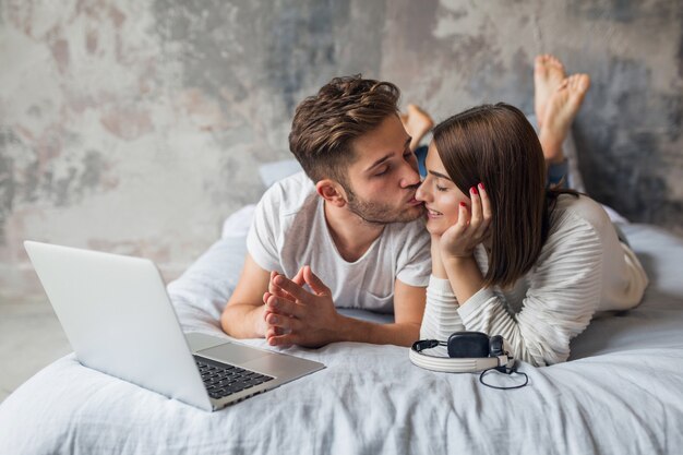 Junges lächelndes Paar, das zu Hause im lässigen Outfit auf Bett liegt und in Laptop, Mann und Frau schaut, die glückliche Zeit zusammen verbringen, entspannend